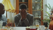 Young joyous Black man sitting at dinner table with eyes closed, blowing candles on cake, smiling and hugging little son during birthday celebration at home
