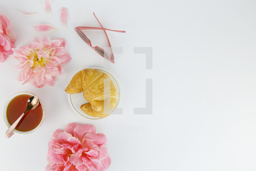 tea cup, pink spring flowers, croissant, sunglasses, and spoon against a white background 
