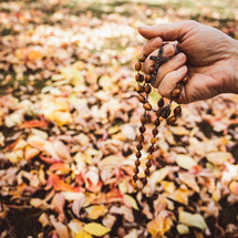 Hand holding a rosary outdoors with ground covered with autumn leaves in background