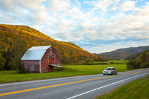 Minivan car driving on scenic Vermont road passing red barn and fall foliage mountains during autumn 