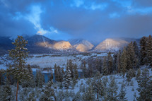 Cloudy snowy winter mountain range landscape with evergreen trees and lake
