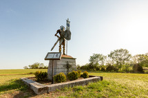 Statue of knight in metal armor on stone column holding sword and shield with sky background
