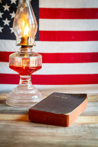 Open Holy Bible and old fashioned oil lamp with American flag background vertical