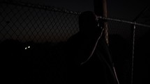 A man standing outside by fence at night.
