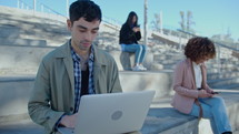 Young handsome businessman sitting outdoors at urban public space in the city and working on laptop, people using phones and walking in the background
