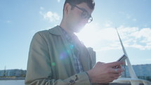 Young businessman standing outside on the riverbank in the city, typing message on mobile phone. Medium shot, low angle view
