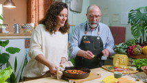 Cheerful senior food bloggers enjoying the smell of hot dish in cooking pan, talking on camera and grating cheese over it while giving online culinary class
