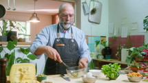 Senior male food blogger in apron cracking eggs in glass bowl, whisking and commenting on camera, filming video recipe or cooking class in the kitchen
