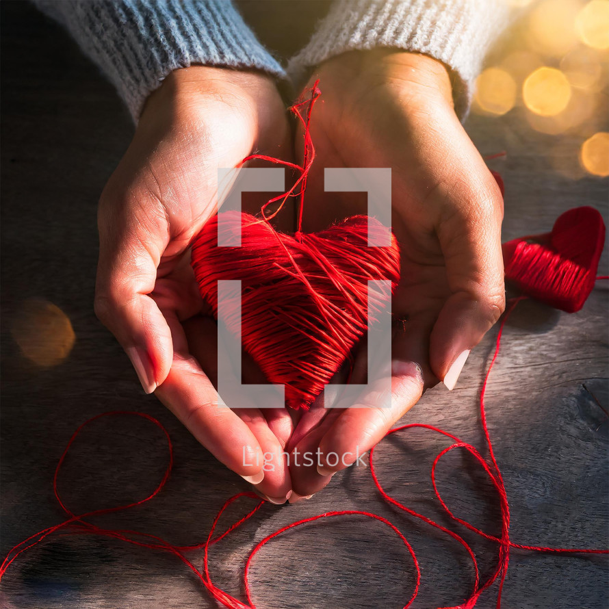 A woman's hands holding a hand-crafted red heart made of yarn or twine on a wooden table for Valentines' Day.