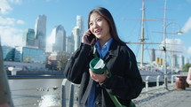 Young positive Asian woman holding coffee travel mug, walking along urban riverside in the city at daytime, smiling and speaking on mobile phone
