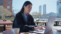 Young Asian businesswoman sitting at table with cup of coffee and croissant, typing on laptop, working remotely in outdoor cafe. Zoom shot, cityscape in the background
