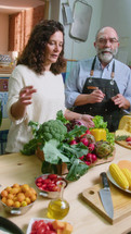 Senior family couple recording video recipe for food blog in the kitchen, telling about cheese and fresh vegetables, showing chefs kiss gesture on camera. Vertical clip
