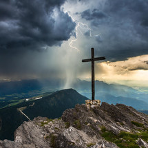 A wooden cross on a mountaintop with a lighting storm in the background and dramatic clouds.