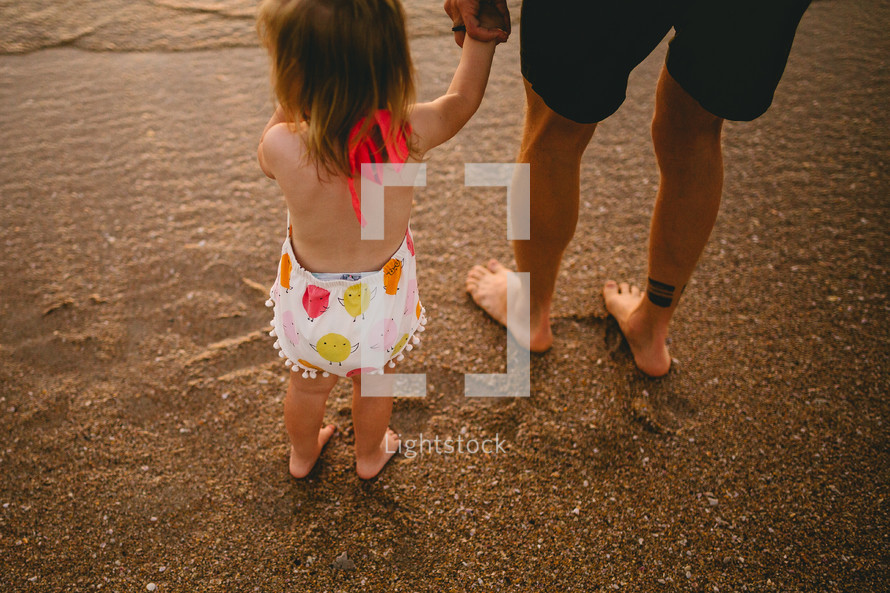 a father and toddler daughter holding hands on a beach 