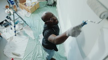 Black man standing on the floor covered with cellophane and painting a wall with roller during DIY apartment renovation. High angle shot
