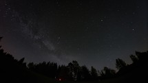 Time lapse of stars, including the Milky Way, move above a silhouette forest. Also satellites and meteors streak across sky. 