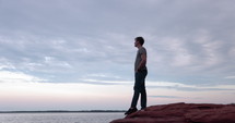 Man standing on edge of rocks looking out into the ocean - wide shot