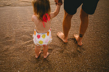 a father and toddler daughter holding hands on a beach 