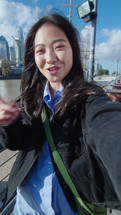 Young beautiful Asian girl standing outdoors on the riverside in the city and speaking on camera, filming blog or video calling. Vertical POV shot
