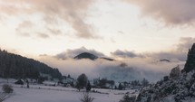 Static Shot of Snowscape Countryside With Tranquil Village Near Forested Mountain Under Overcast Sky During Sunset. 