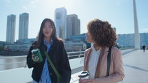 Young Asian woman holding a coffee mug and talking with African-American female friend while walking outdoors in the city after workday. Medium shot
