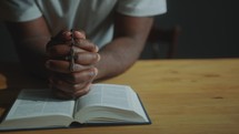 Hands of Black Man Holding Rosary Cross and Praying with Bible
