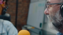 Senior podcast host with gray beard and headphones talking into microphone with colleague in a recording studio. Close-up view