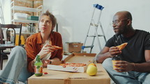 Young married couple sitting on the floor in living room, eating pizza, having drinks and talking when taking a break during home renovation. Zoom-out shot
