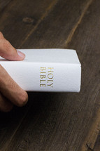 A hand holding a white Bible on a wooden table.