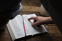 A man's hand pointing to a verse in an open Bible.