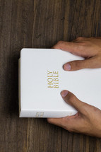 Two hands holding a white Bible on a wooden table.
