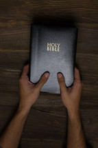 A man's hands holding a black Bible on a wooden table.
