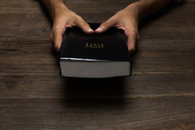 A man's hands holding a black Bible on a wooden table.