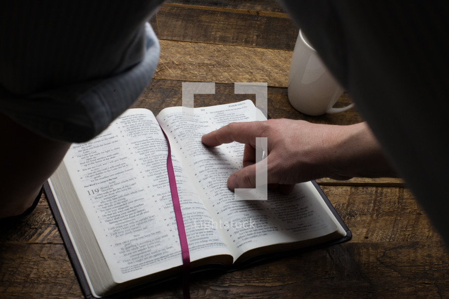 A man's hand pointing to a verse in an open Bible.