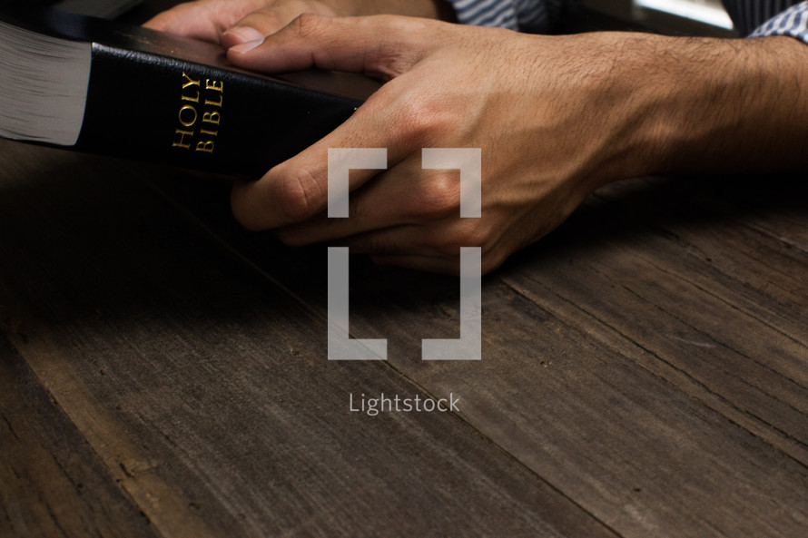 Hands holding onto a Bible on a wooden table.