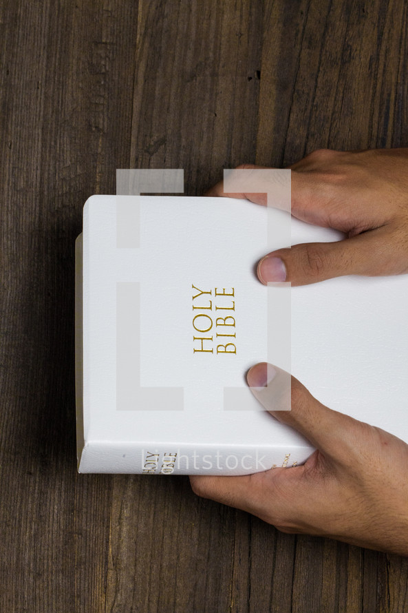 Two hands holding a white Bible on a wooden table.