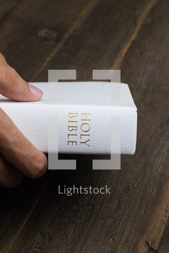 A hand holding a white Bible on a wooden table.
