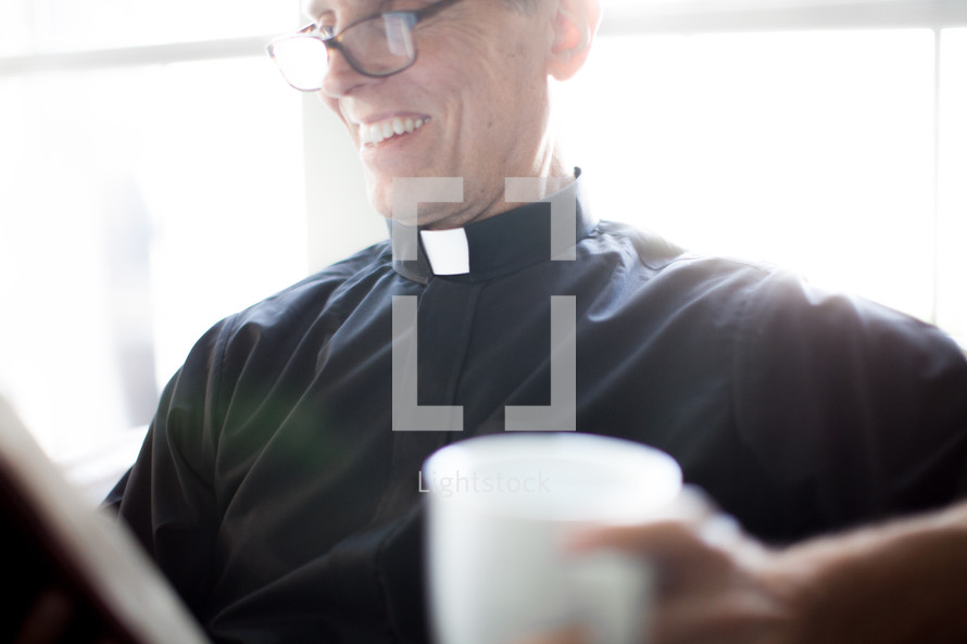 A smiling minister in clerical clothes reads the Bible and holds a cup of coffee.