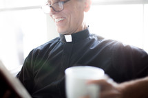 A smiling minister in clerical clothes reads the Bible and holds a cup of coffee.