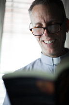 A smiling minister wearing a clerical collar reads the Bible.