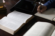 A man's hand takes notes at a desk upon which is an open Bible.