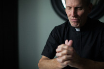 A minister wearing clerical clothes clasps his hands and closes his eyes in prayer.
