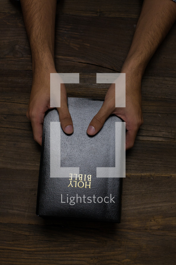Two hands holding a Bible on a wooden table.