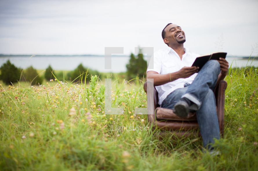 man sitting outdoors in a chair in a field reading a Bible 