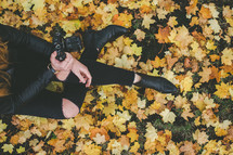 A woman with a camera sitting in fall leaves.
