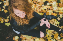 A woman with a camera sits on the ground among fall leaves.