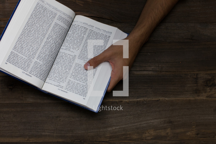 A hand holding an open bible on a wooden table.