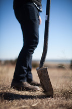 A man with a shovel stands in a field of brown grass.