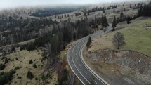 Aerial shot of a highway on sloping hills with conifer forest trees against a moody sky.