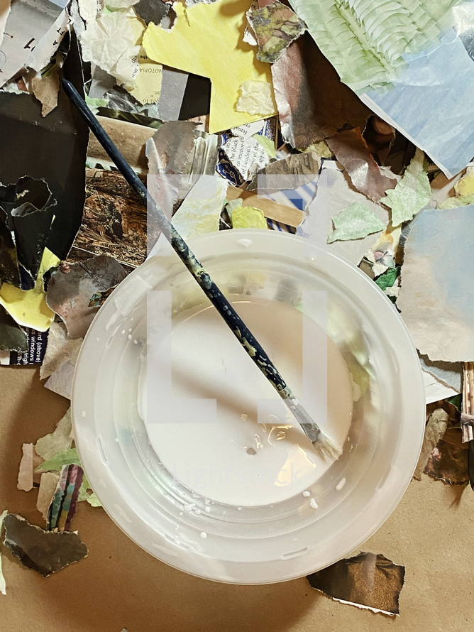 artist's brush resting in glue bowl with torn and cut collage papers on work table